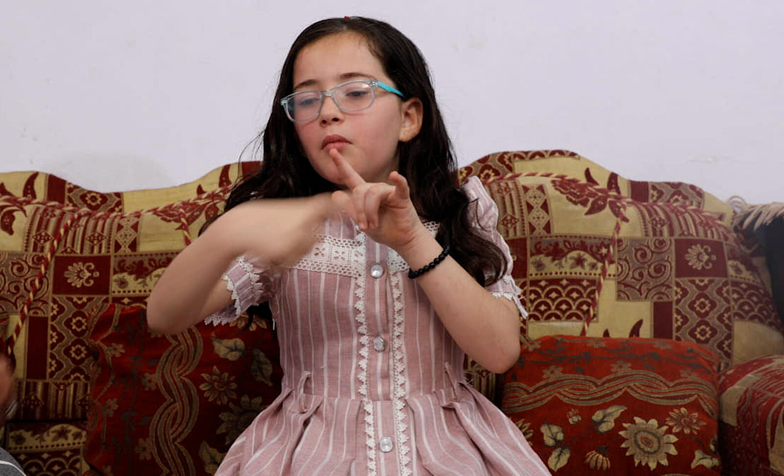 A girl in a pink dress signs, one finger raised.